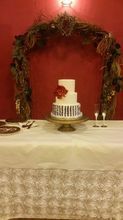 Three-tiered wedding cake with red flower on a pedestal table, beneath a rustic arch against a red wall.