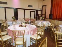 Banquet hall with round and rectangular tables set for a celebration. Gold chairs, pink and white linens.