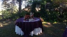 Table set for an outdoor event with purple tablecloth over a white one, in a natural setting.