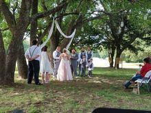 Wedding ceremony under trees. Bride and groom with wedding party, decorations and guests on a grassy area.