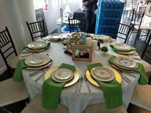 Round table set for a formal event with white tablecloth, gold chargers, green napkins, and black chairs.