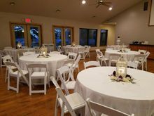 Reception hall set for a party, with round tables draped in white, white chairs, and decorative lanterns.