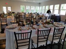 A decorated dining hall with round tables set for an event; black chairs; windows with outdoor views; someone at a table.