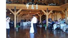 Couple dances at wedding reception in a rustic barn with string lights, tables, and guests.