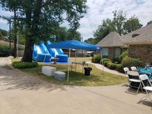 Outdoor party scene with blue water slides, tent, tables, chairs, and a brick house.