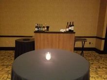 A bar with bottles and glasses, a round table with a candle, and a black trash can.