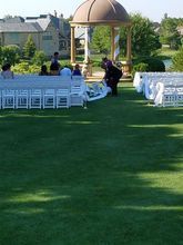 Wedding preparations: People setting up chairs and decorations on a green lawn near a gazebo and houses.