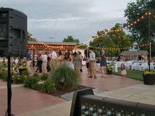 Outdoor event: People gathered on a brick patio. String lights overhead. Speaker in foreground. Cloudy sky.