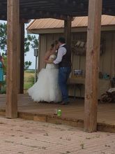 Bride and groom embrace on wooden deck; rustic setting.