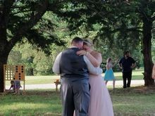 Bride and groom dance outside; she wears a pink dress, he a gray vest. Guests watch.