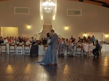 Couple dancing in a wedding reception hall, guests seated at long tables, crystal chandelier overhead.