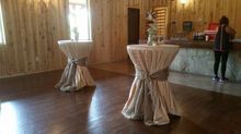 Two cocktail tables draped with tan fabric and brown sashes in a wood-paneled room; person at a bar.
