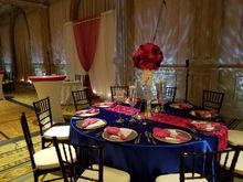Round table set for a formal event with blue tablecloth, magenta runner, and floral centerpiece in a grand hall.