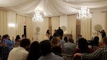 Elegant event space with chandeliers and draped ceiling. Guests seated, couple at the front.