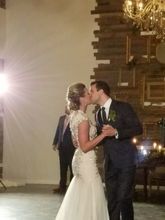 Bride and groom kiss during a wedding dance. Bride in white dress, groom in suit, in a venue with a wall of wood.