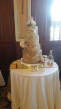 Four-tiered gold cake with floral accents sits on a white-covered table.