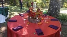 Round table set for a fall event with red tablecloth, purple napkins, and floral centerpiece in a wooded area.