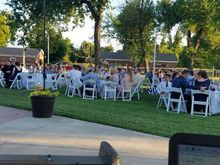 Outdoor event with guests at tables on grass. White chairs and tablecloths. Trees and buildings in the background.