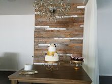 Cake table with two-tiered cake, cupcakes, against a wood-paneled wall, under a chandelier.