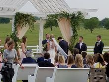 Wedding ceremony under an arbor, bride and groom exchanging vows, guests seated on white benches, outdoor setting.