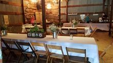 Tables set for a rustic event inside a barn, with chairs, floral centerpieces, and food service in the background.