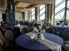 Wedding reception setup: round tables with navy blue tablecloths, silver runners, and centerpieces in a room with large windows overlooking the view.
