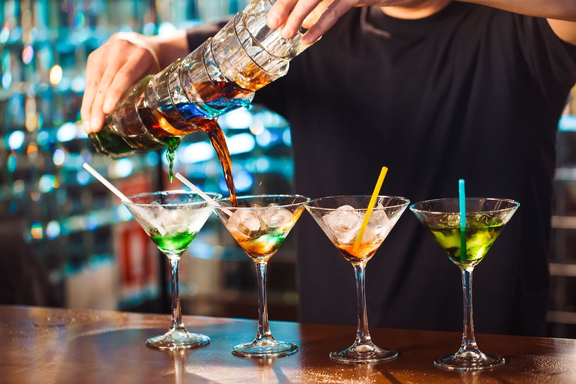 Bartender pouring a layered drink into colorful cocktails with ice and straws.