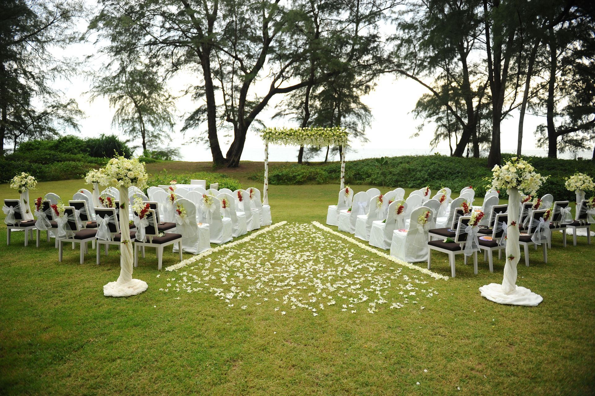 Wedding ceremony setup on green grass with white chairs, floral aisle, and arch; trees in background.