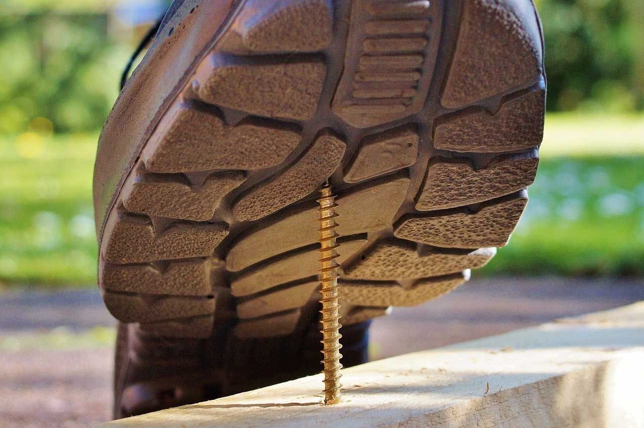 Close-up of a boot stepping on a nail embedded in a concrete curb, with a blurred outdoor background