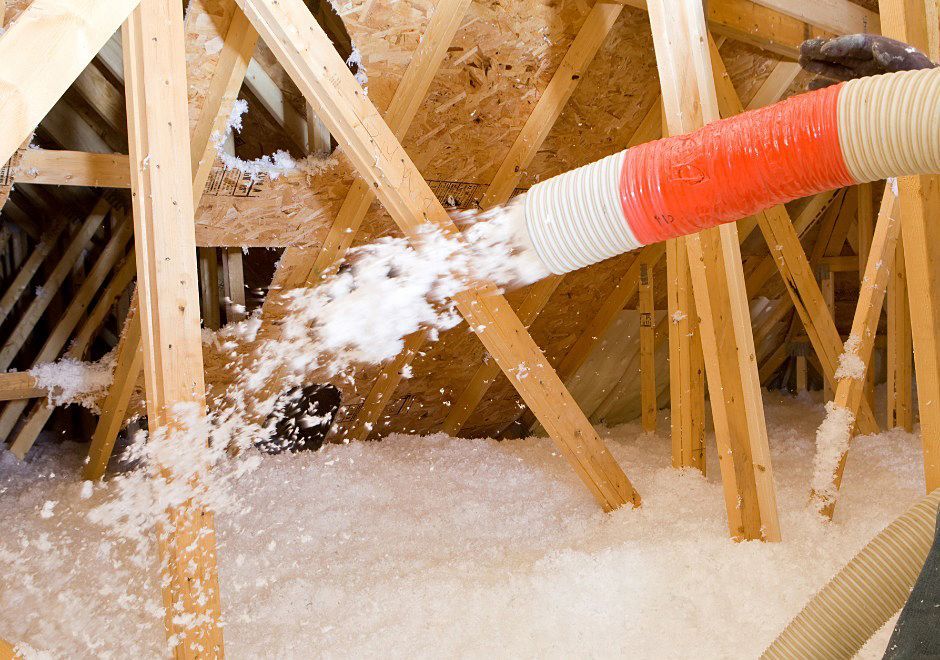 Attic being insulated with white fluffy insulation, sprayed from an orange and white hose, between wooden rafters.