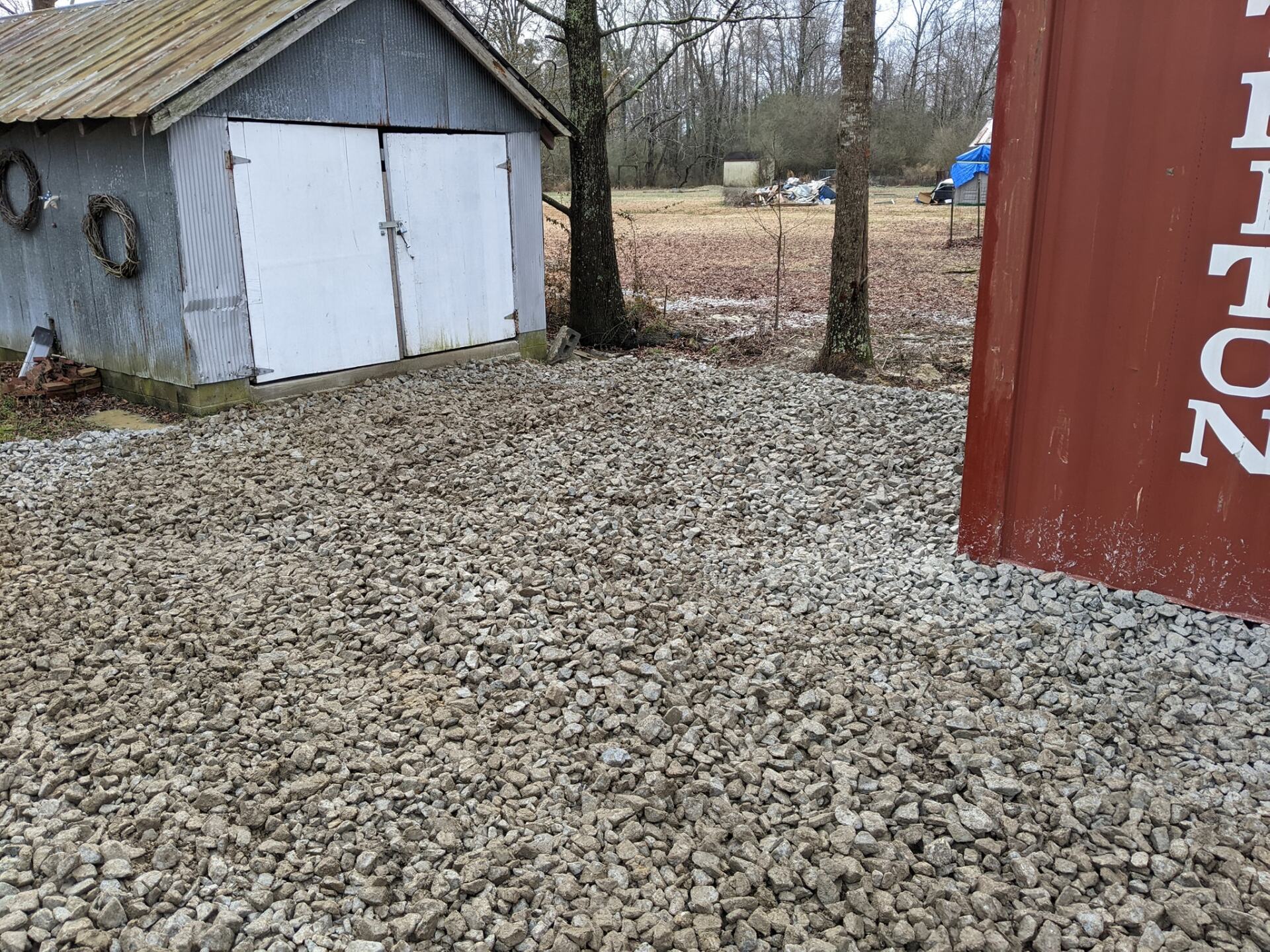 a shed is sitting in the middle of a gravel driveway next to a red container .