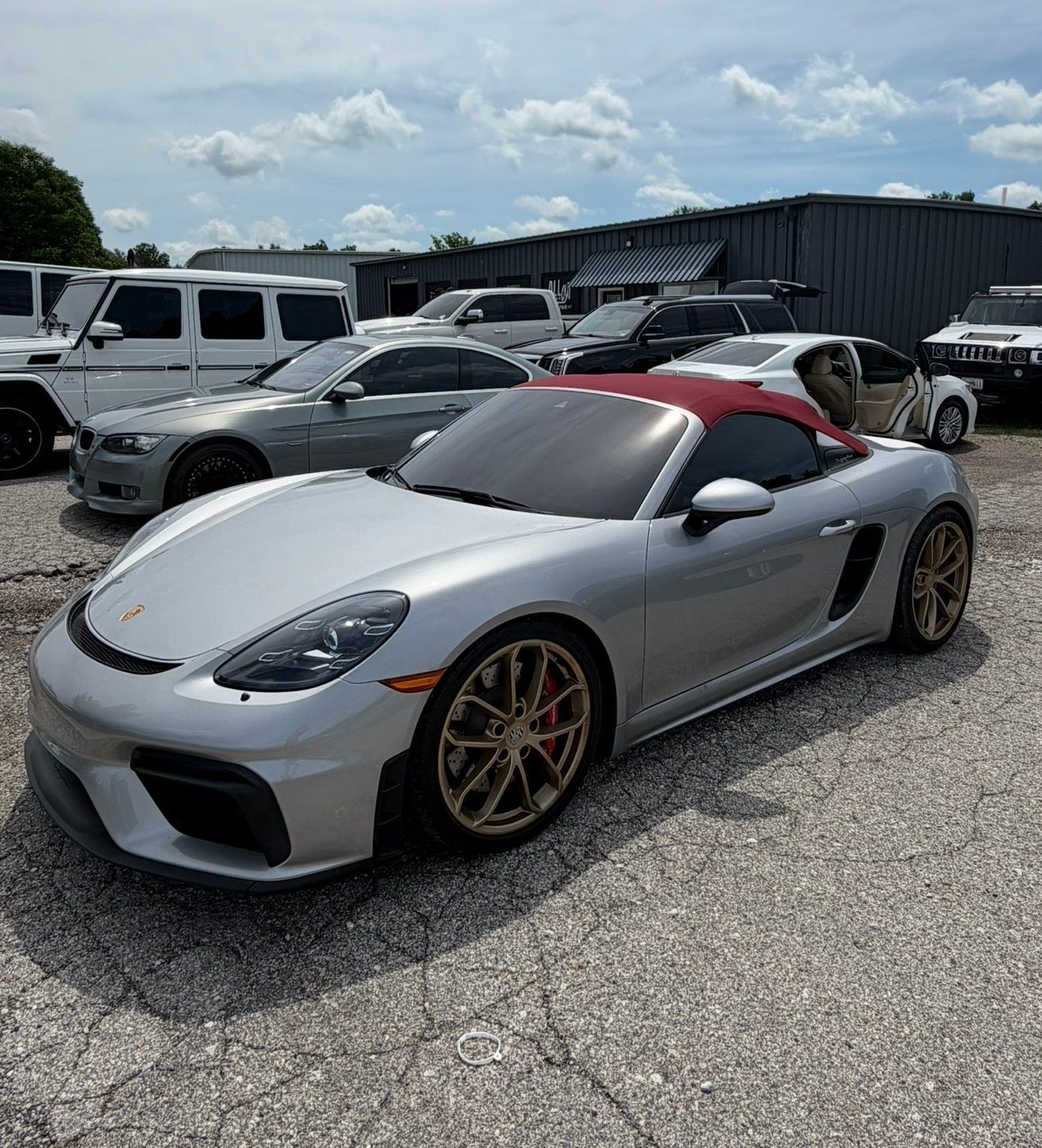 Silver sports car with red roof and gold rims parked in a gravel lot under a cloudy sky