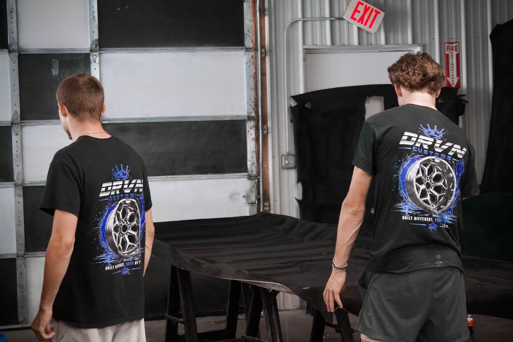Two people in black shirts moving a covered table inside a garage-like workspace