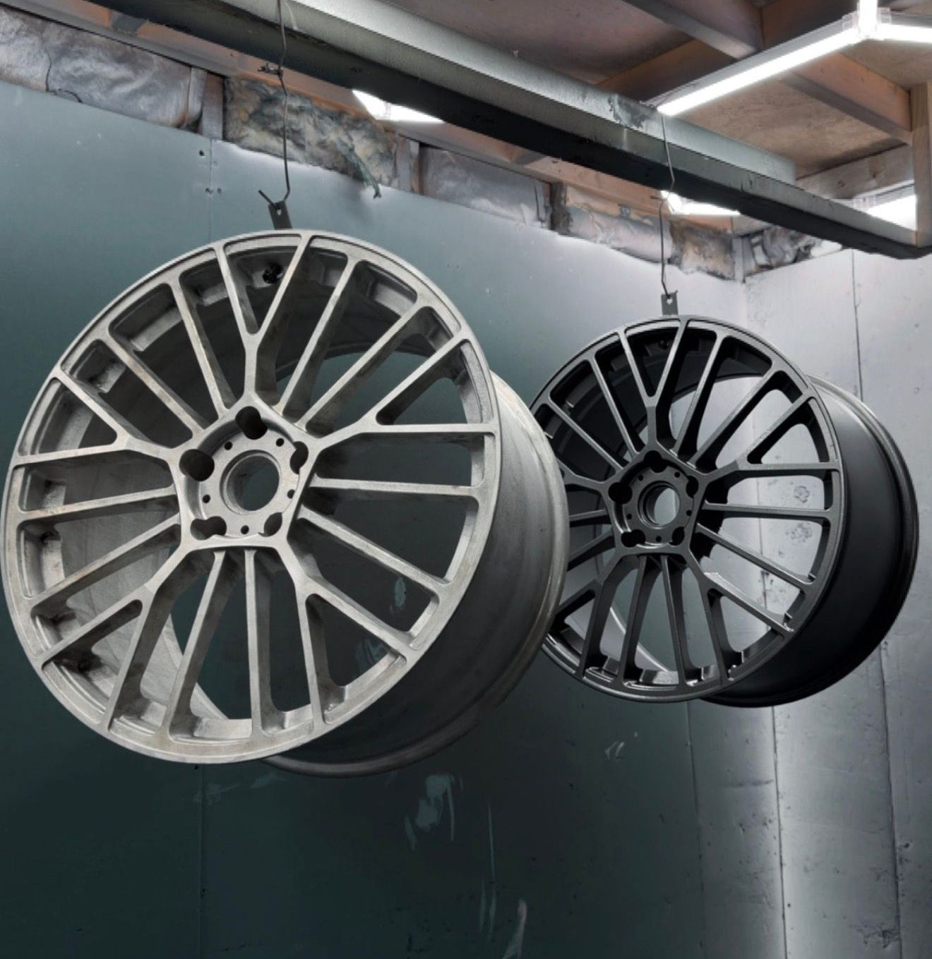 Two alloy car wheels, one silver and one black, hanging in a workshop spray booth.