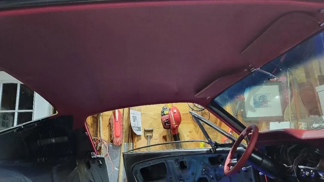 Inside a car with maroon ceiling, steering wheel, and yellow street scene visible through the windshield