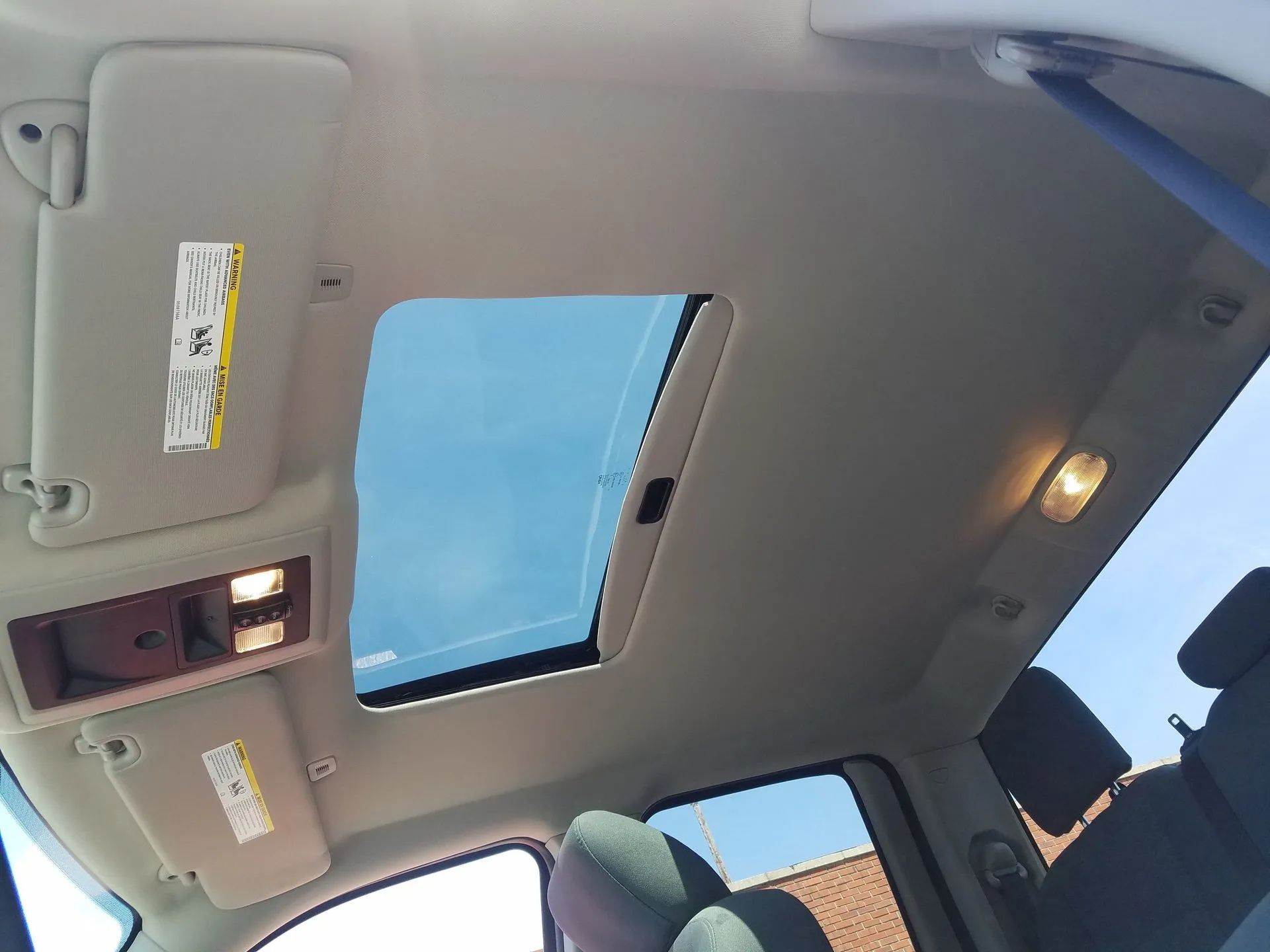 Car interior ceiling with sunroof open, dome light, visors, and a view of blue sky above.