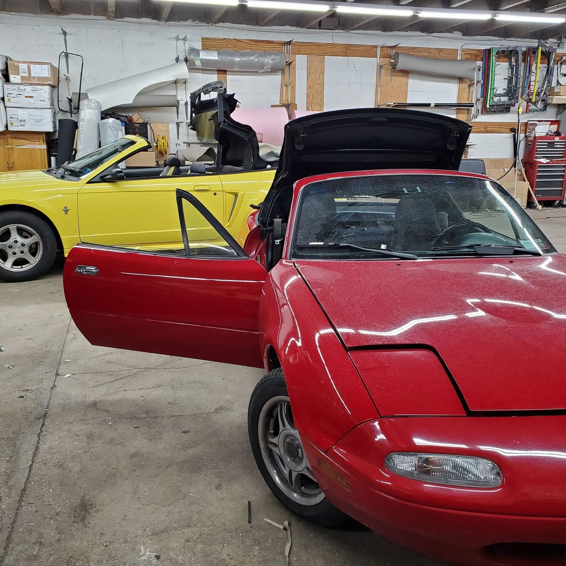 Red convertible with open door and trunk in a garage, with a yellow sports car in the background