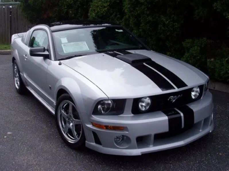 Silver sports car with black racing stripes parked on a road near trees