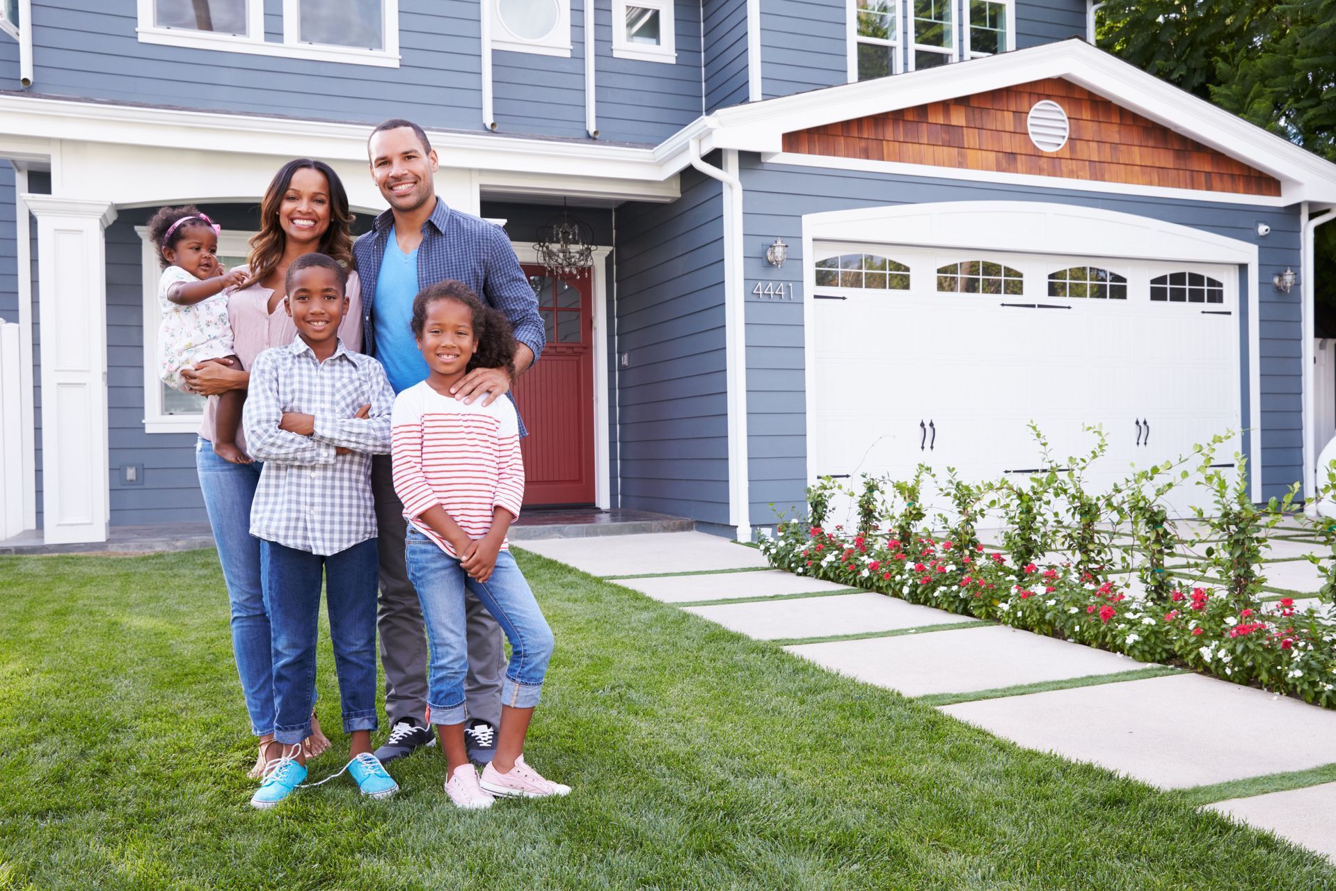 Family of five smiling in front of their blue house with green lawn.
