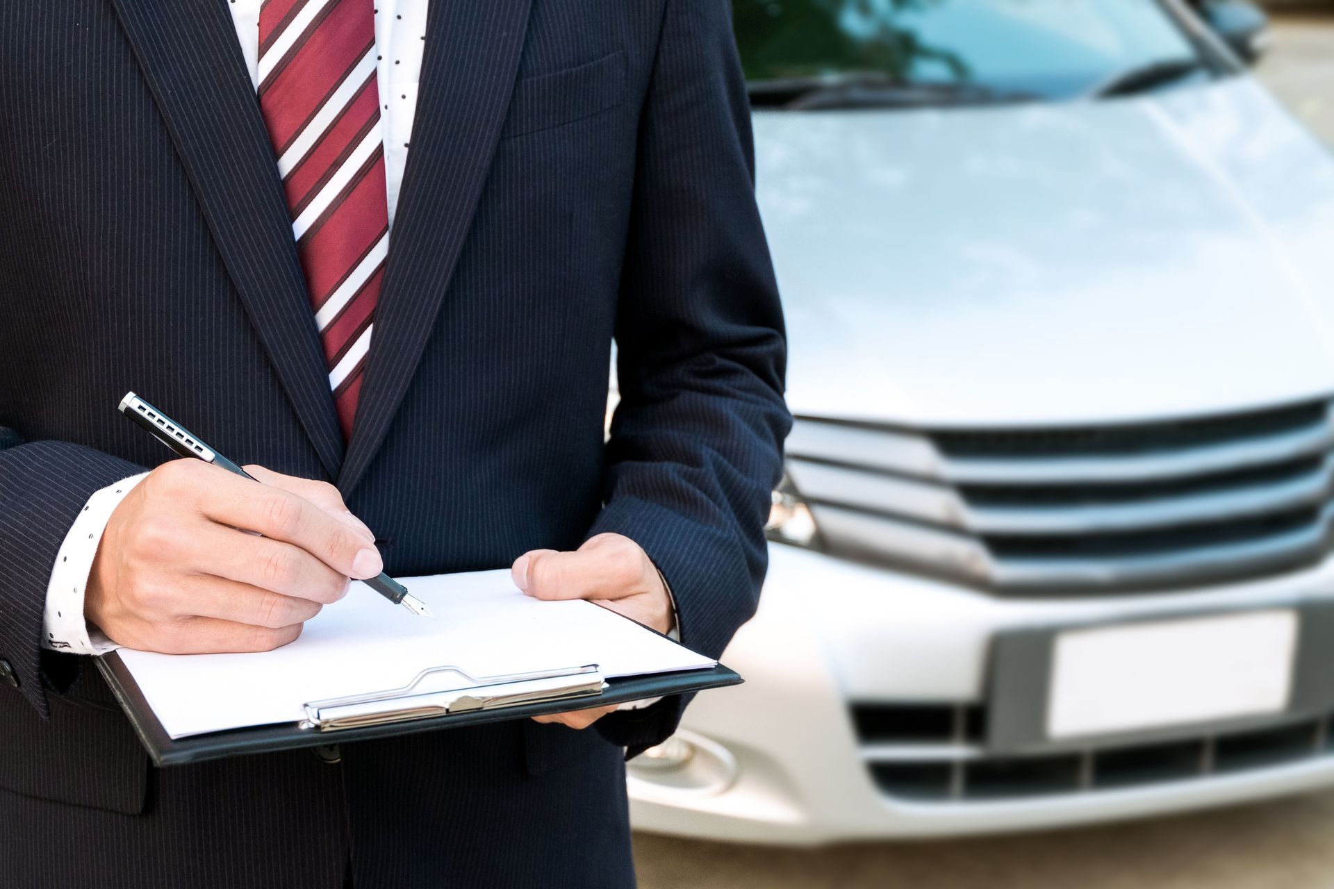 Person in suit writing on clipboard next to a silver car.