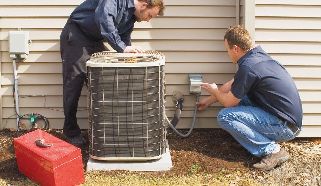 Two HVAC technicians servicing an outdoor air conditioning unit next to a beige house. One kneels, working on electrical.