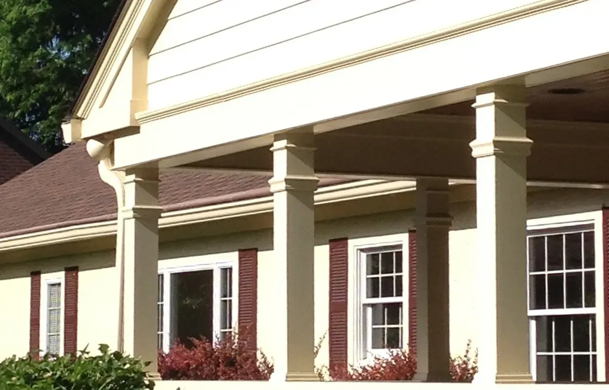 Cream-colored house porch with white columns, red shutters, and burgundy shrubs.
