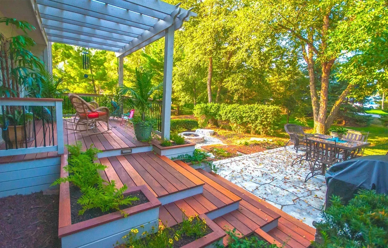 Covered backyard deck with red wood steps, wicker seating, and a stone patio surrounded by lush greenery.