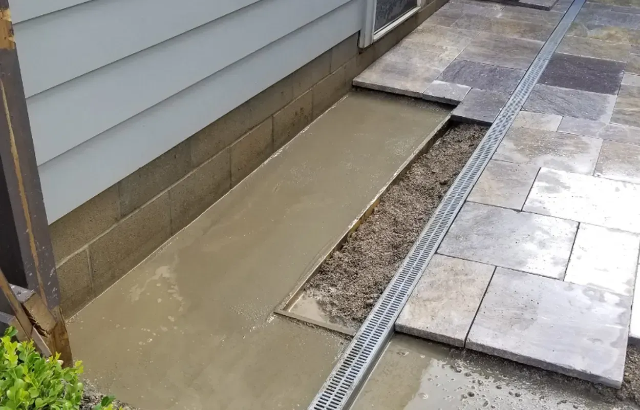 Outdoor side yard with wet concrete strip beside a house, pavers, gravel, and a drainage channel