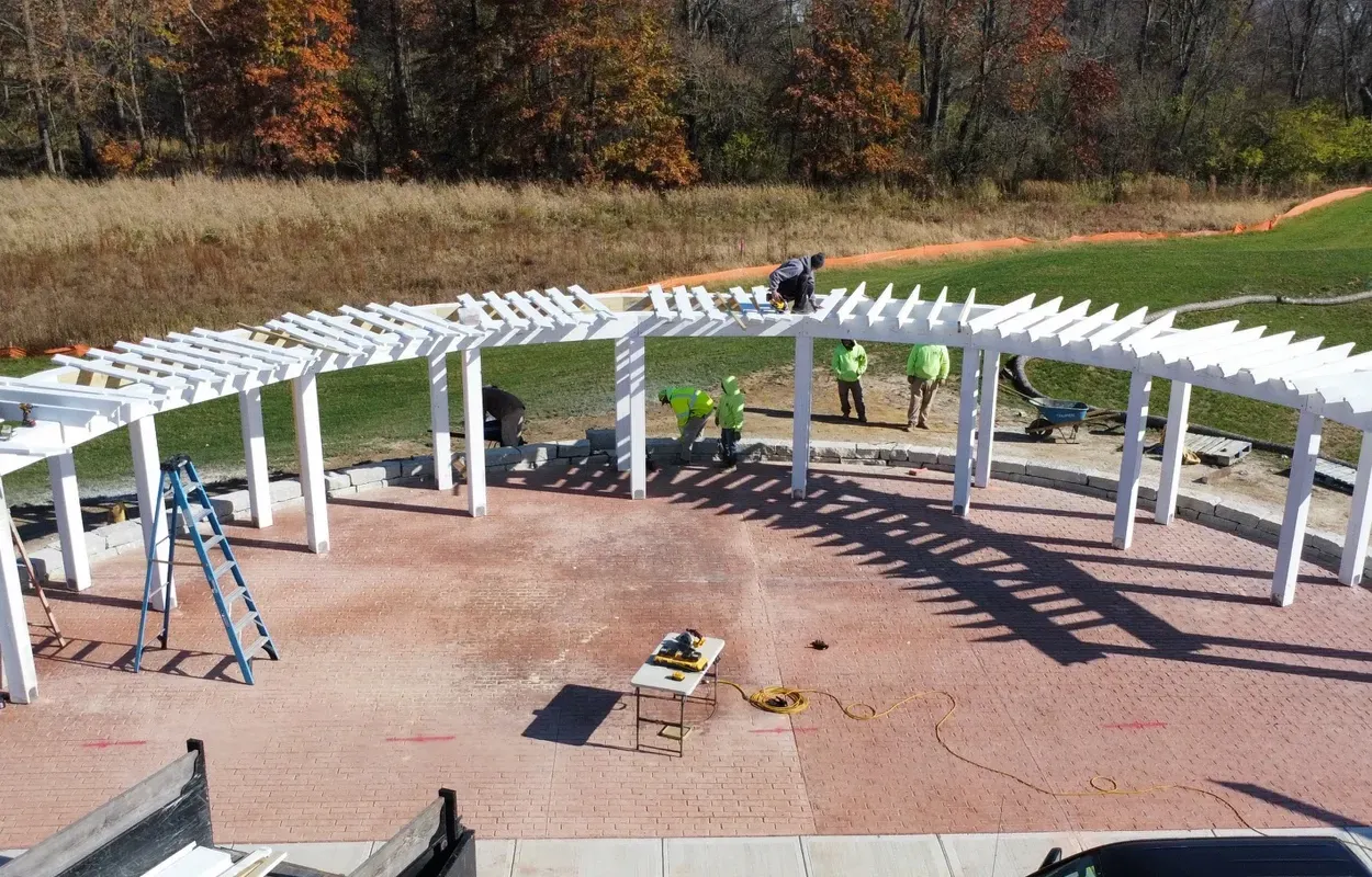 Outdoor construction of a white pergola structure with ladders on a brick patio, workers installing beams