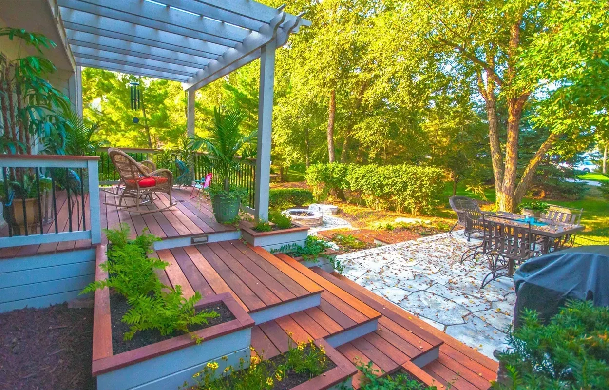 Backyard deck with pergola, wooden steps, potted plants, and a stone patio surrounded by trees