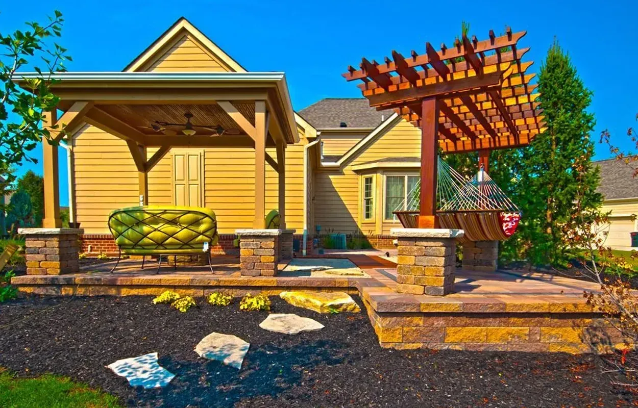 Two wooden pergolas in a landscaped backyard with stone steps, mulch, and a house in the background