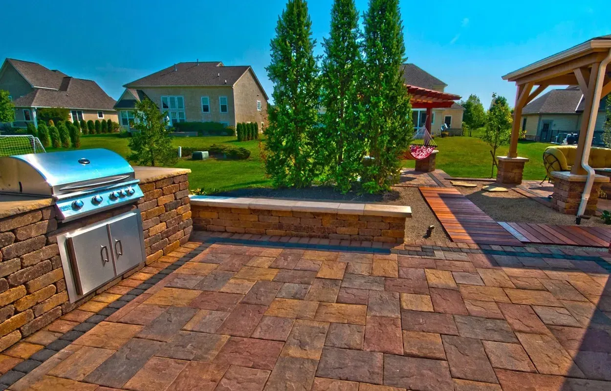 Backyard patio with stone grill, built-in bench, pavers, and landscaped lawn under a clear sky