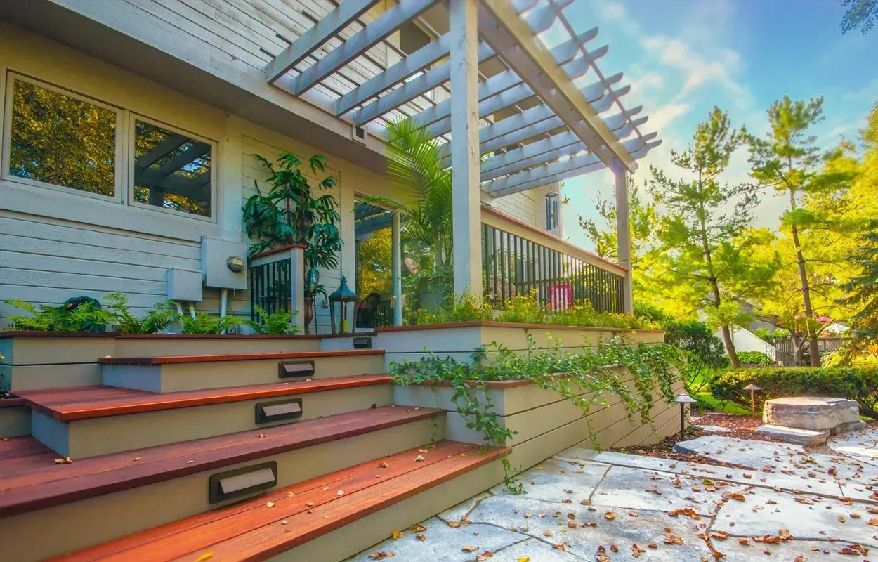 Front steps to a white house with a pergola, potted plants, and sunlit trees in the yard