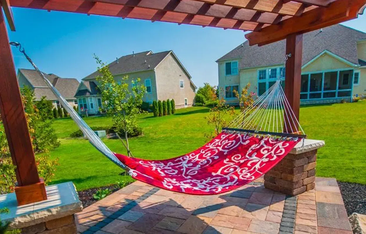 Red hammock on a covered patio overlooking a sunny suburban backyard with houses and green lawn.